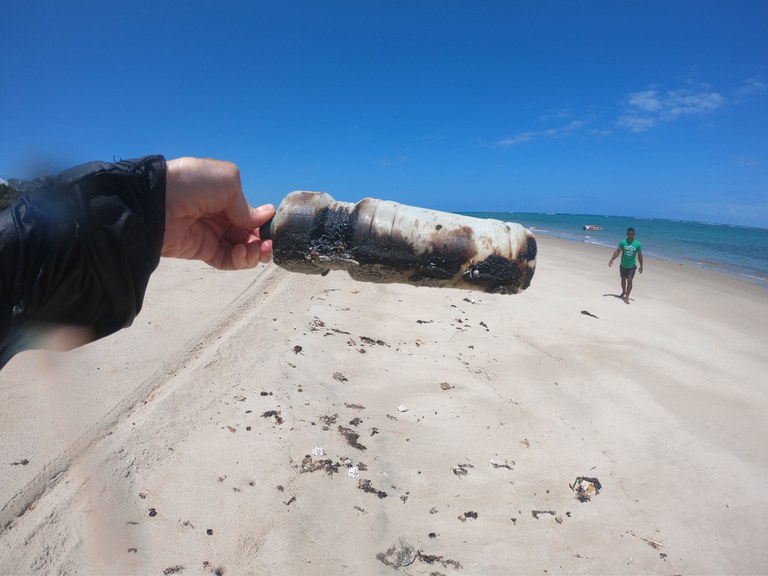 mão em primeiro plano segurando garrafa de plástica com resíduos de óleo e praia em segundo plano
